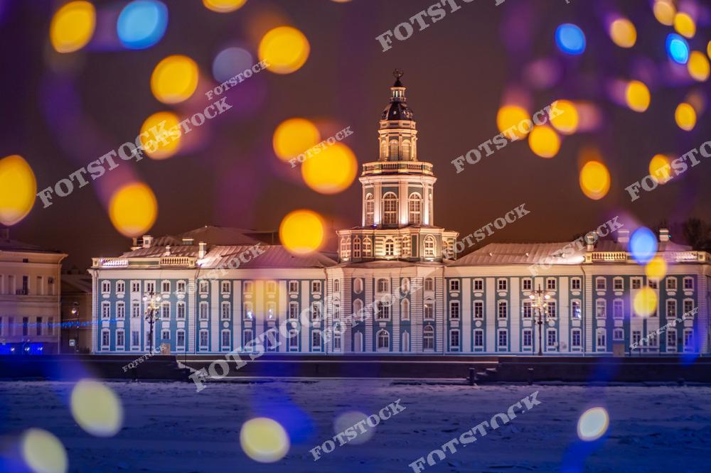 На фотографии изображена Кунсткамера — одно из старейших зданий Санкт-Петербурга, подсвеченное ночью. Яркие огни переднего плана создают ощущение праздничности и подчёркивают историческую значимость музея, отражающего научное и культурное наследие города. Снежная набережная и мягкое ночное освещение добавляют уют и волшебство в зимний пейзаж Петербурга, привлекая внимание к уникальной архитектуре здания.
